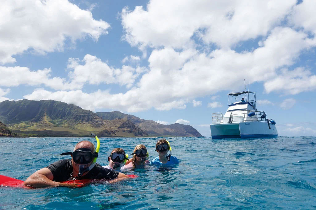 A family snorkeling together beside the Hale Kai catamaran below the green Waianae mountains